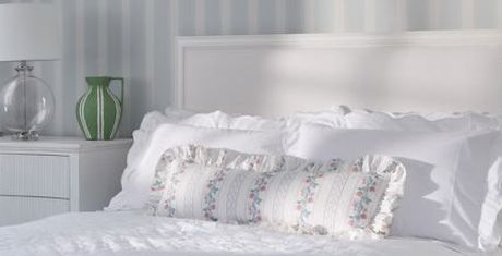 A neatly made bed with white linens and floral pillows, next to a nightstand with a green vase and a lamp. Striped wallpaper in the background.