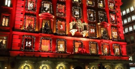 A building facade illuminated in red with numbered windows, resembling an advent calendar, decorated with festive lights and wreaths.
