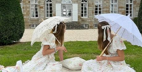 Two women in floral dresses sit on grass with lace parasols, facing a historic stone building with arched windows and white shutters.