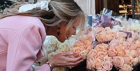 Woman in a pink coat smelling a bouquet of peach roses outside a flower shop.