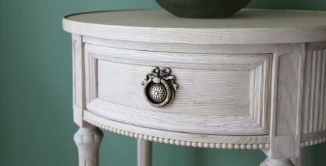 Round wooden side table with ornate drawer handle and a green vase on top, set against a green wall.
