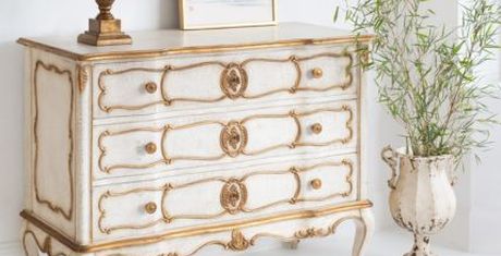 Elegant white and gold vintage dresser with ornate details, a framed picture, a decorative vase, and a potted plant on a white background.