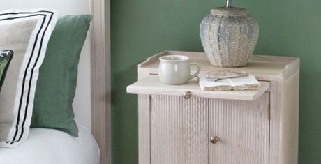A wooden nightstand with a ceramic lamp, a mug, and an open book. It is next to a bed with green and white pillows.