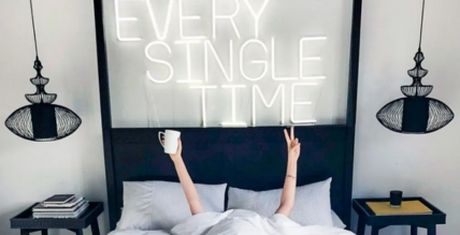 Person in bed holding a mug and making a peace sign, with neon sign saying "EVERY SINGLE TIME" above. Modern lamps and side tables flank the bed.
