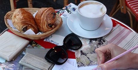 A table with two croissants in a basket, a cup of coffee, sunglasses, and a person writing with a pencil on paper.