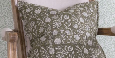 Green cushion with intricate white floral patterns on a wooden chair, set against a pale patterned background.