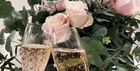 Two champagne flutes clink in front of a bouquet of pink roses and green leaves, creating a celebratory and romantic atmosphere.
