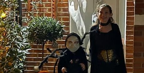 Two children in Halloween costumes stand outside a decorated house. One wears a mask and spider costume, the other in a dark outfit with a gold belt.