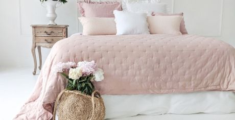 A bed with a pink quilt and assorted pillows, beside a wooden nightstand with a vase of flowers and a woven basket on the floor.