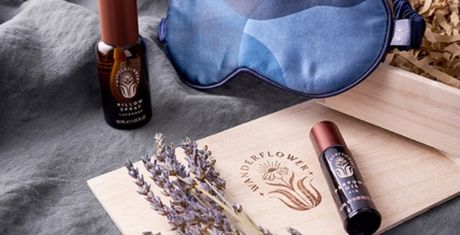 A sleep mask, lavender sprigs, and two essential oil bottles on a wooden box with shredded paper, all arranged on a soft fabric.