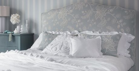Elegant bedroom with a floral headboard, white bedding, decorative pillows, and a blue nightstand with a lamp and flowers. Striped wallpaper background.