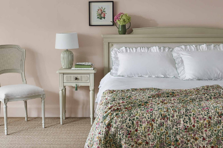 A cozy bedroom features a cream bed with white pillows and a floral quilt, a matching beige nightstand with a lamp and vase, and a chair beside it.