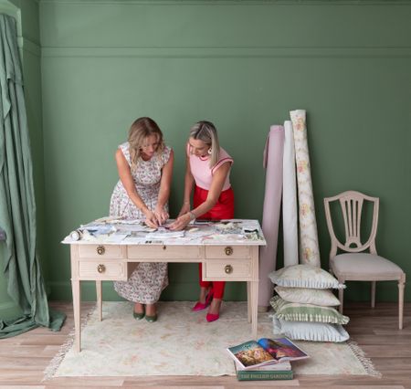 Two women examining fabric samples on a table in a green room, with rolls of fabric and cushions nearby.