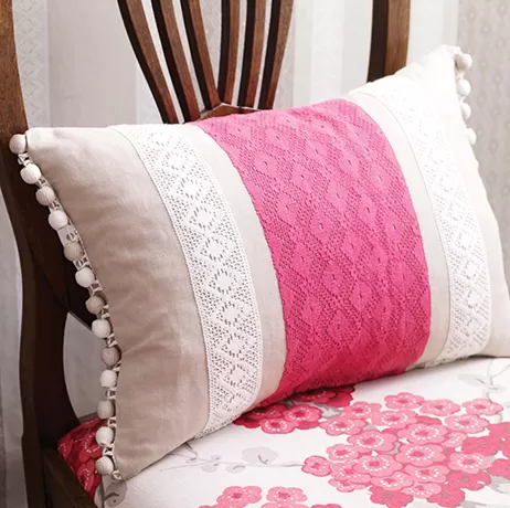 A decorative pillow with pink lace and white fabric, adorned with pom-poms, rests on a wooden chair with a floral-patterned seat cushion.