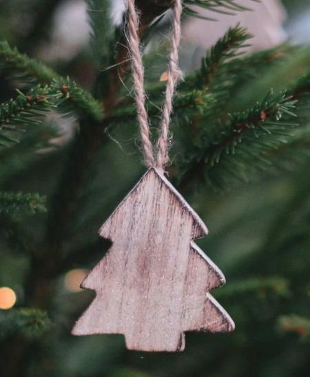 Wooden Christmas tree ornament hangs on a green pine branch, with soft-focus lights in the background.
