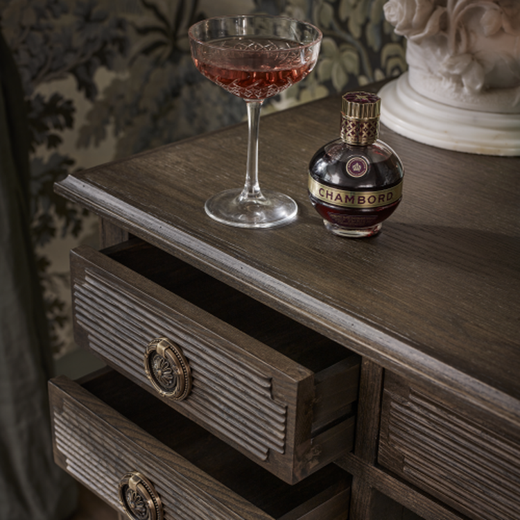 A glass of red liquid and a Chambord bottle sit on a wooden dresser with open drawers, against a floral-patterned wallpaper.