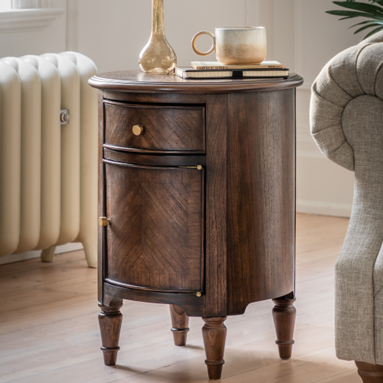 Wooden round side table with brass handles, a gold vase, books, and a mug on top, next to a radiator and a beige sofa on a wooden floor.