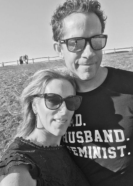 A couple wearing sunglasses poses outdoors. The man wears a shirt with text, and the woman has long hair. A fence and people are in the background.