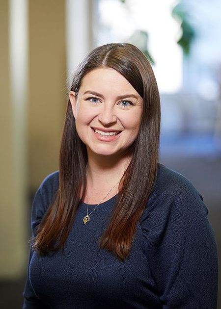 Smiling woman with long brown hair, wearing a dark blue top, stands in a softly lit room with a blurred background.