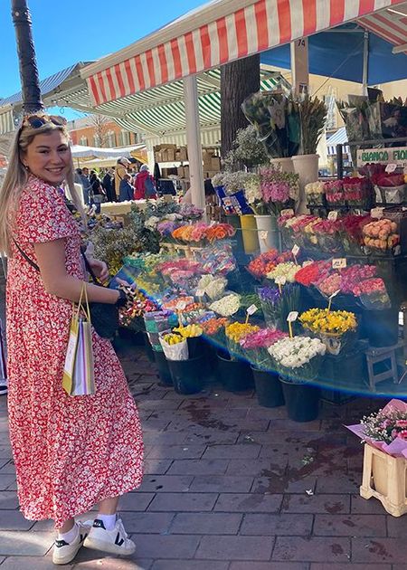 Woman in a red floral dress smiles while holding shopping bags at an outdoor flower market with various colorful bouquets.