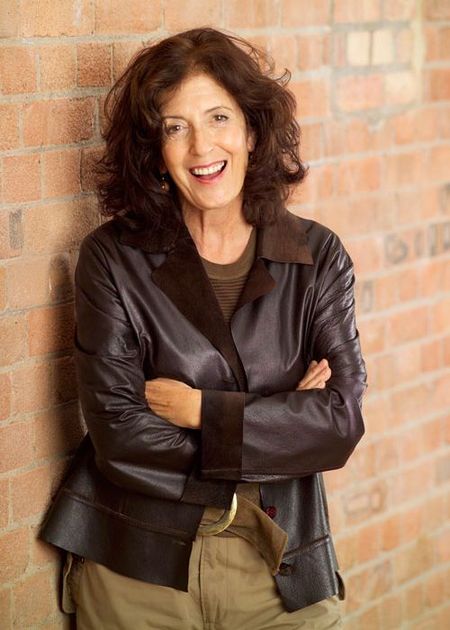 Smiling woman with curly hair in a leather jacket stands against a brick wall, arms crossed.