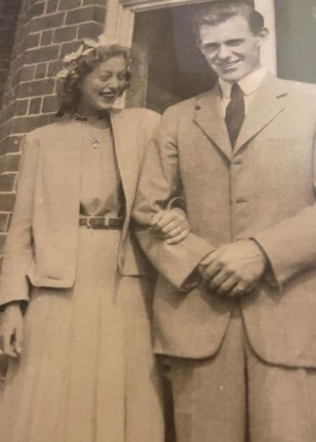 Vintage photo of a smiling couple in formal attire, standing arm in arm outside a brick building.