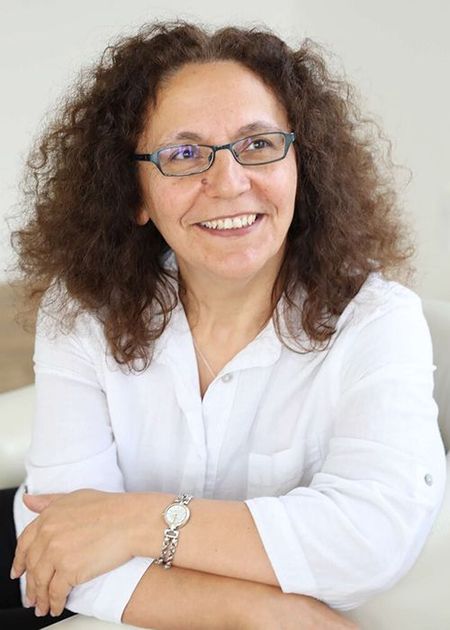 Smiling woman with curly hair and glasses, wearing a white shirt and a silver watch, seated with arms crossed on a white surface.