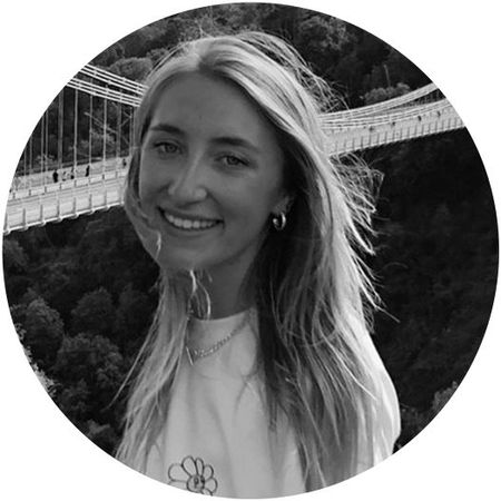 Smiling person with long hair stands in front of a suspension bridge, with a forested background. Black and white image.