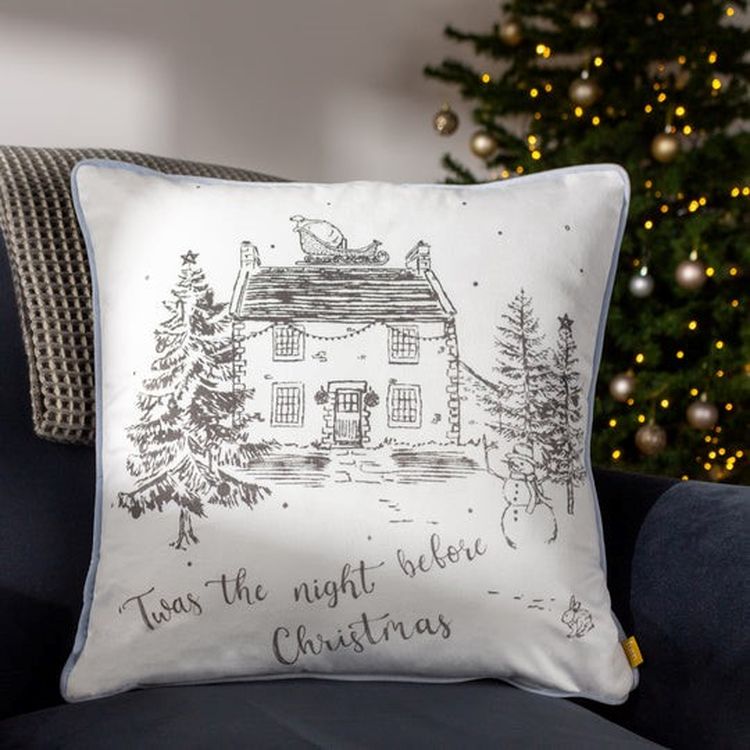 Festive pillow with a sketch of a house, trees, and snowman, inscribed with "Twas the night before Christmas," near a decorated Christmas tree.
