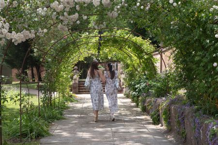 Two woman in toile dresses walking under a rose arch in the Glyndebourne grounds.