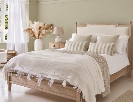 A cosy bedroom with an upholstered wooden bed, white and green striped pillows, and a beige throw in a pale green painted room.