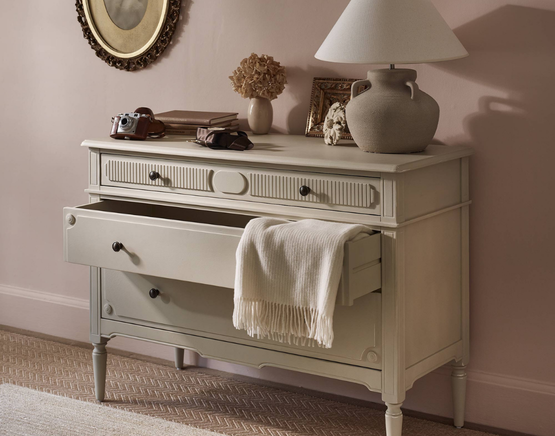 A stone cooured vintage-style dresser with an open drawer draped with a white cloth, topped with a lamp, books, camera, vase, and photo frame against a pink wall.
