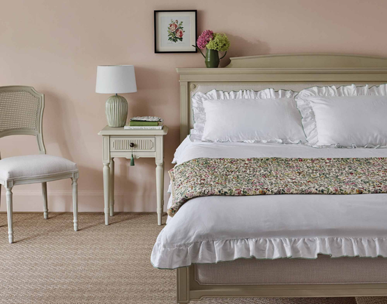 Elegant bedroom with a cream upholstered bed, matching cream bedside table, frilled white bedding, floral quilt, green lamp, and floral artwork on a soft pink wall, accompanied by a side chair.