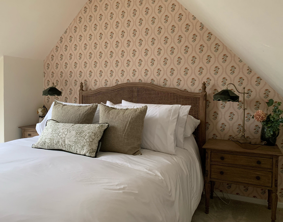 Cosy slanted ceiling bedroom with a rattan bed featuring white linens and decorative pillows, floral wallpaper, and wooden nightstands with green lamps.