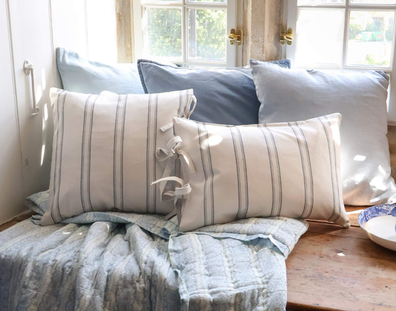 Blue and white stripe cushions sit on a weathered wood window seat with a blue floral quilt and various shades of blue linen cushions, besides white furniture and a brightly lit window.