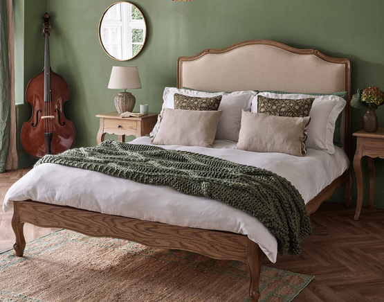 Cozy bedroom with a wooden bed, green wall, decorative pillows, knitted throw, bedside tables, a lamp, plants, mirror, and a double bass.