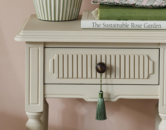 Pale beige bedside table with a drawer, Gustavian Fluting detail, and a round knob with green tassel.