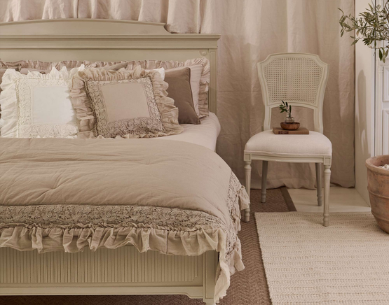 Elegant bedroom with a ruffled beige bedspread, decorative pillows, a vintage chair, and a small plant, creating a cosy, rustic ambiance.