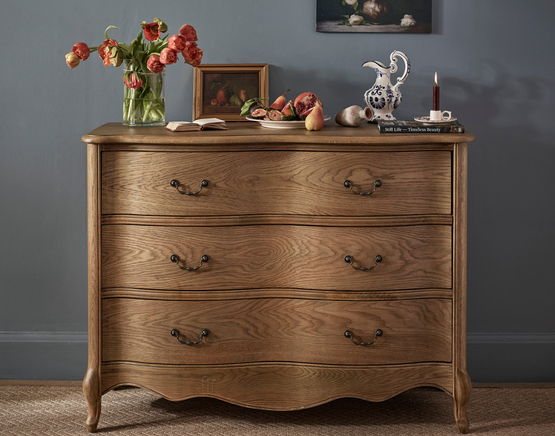 A wooden dresser with ornate handles holds a vase of flowers, open book, fruit, decorative jug, and candle, set against a gray wall.