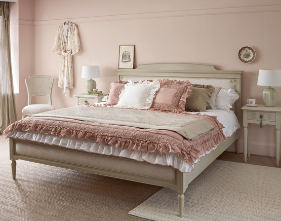 Aa upholstered warm white bed adorned with pink and beige ruffled pillows. Soft pink walls and elegant décor create a serene atmosphere.