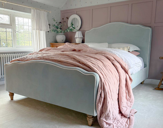 A cosy bedroom with a pale blue upholstered bed, pink quilt, white pillows, and a soft carpet.