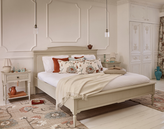 A cream wooden bed styled with rust and blue hints through floral pillows, décor items and a patterned rug, plus a beige blanket, matching bedside tables and glass pendant lights.