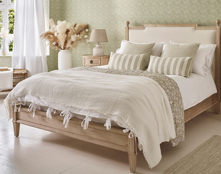 Cozy bedroom with a light wood bed, white and green bedding, striped pillows, and a vase with pampas grass on a light wood floor and patterned green walls.