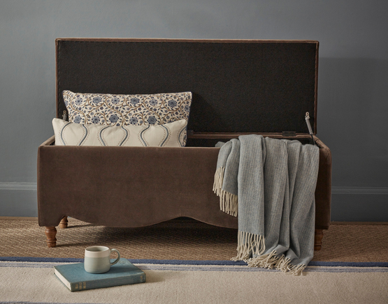 Brown velvet storage bench with open lid, holding patterned cushions and a blue throw. A book and mug sit nearby on the floor.