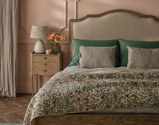 Cosy bedroom with a wooden bed, green and beige cushions, floral quilt, and a nightstand holding a lamp and flowers. Light pink walls.