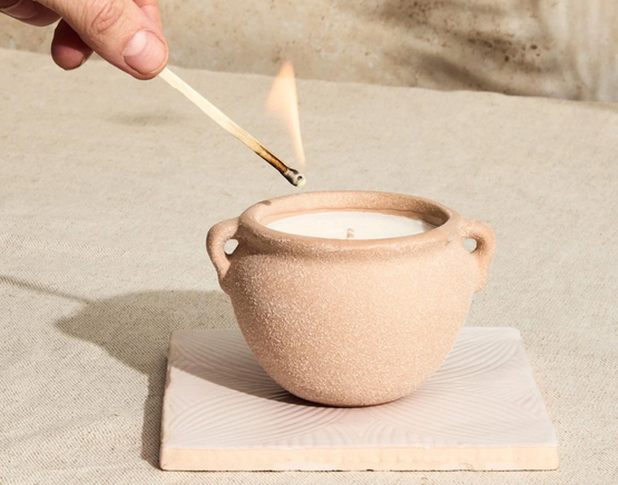 A hand holding a lit match approaches a textured ceramic candle to light it