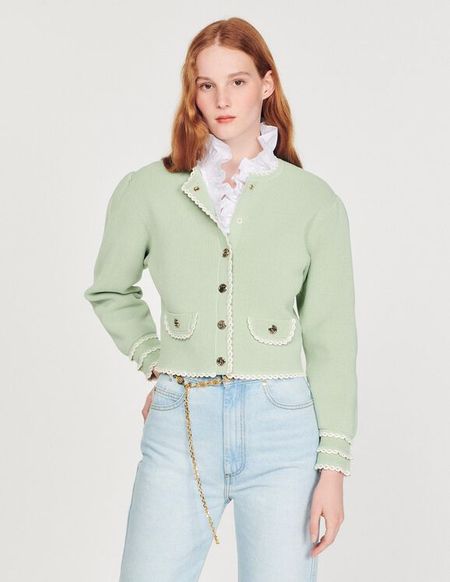A person with long red hair wears a light green cardigan, white ruffled blouse, and light blue jeans, posing against a plain background.