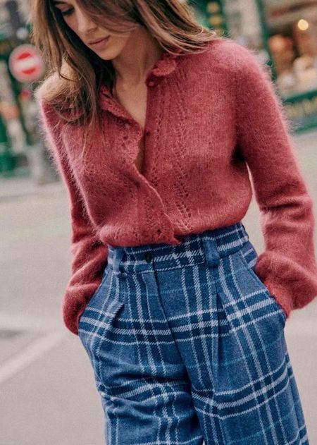 Woman in a pink fuzzy sweater and blue plaid pants walks on a city street, with blurred buildings and a traffic sign in the background.