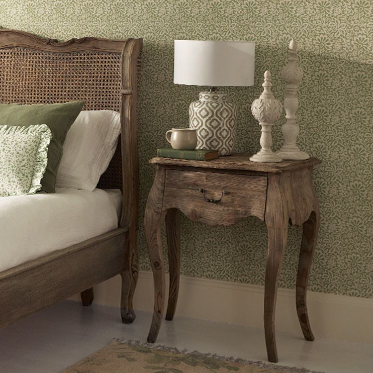 Rustic bedroom with a wooden bed and nightstand, a patterned lamp, decorative objects, and a cup, set against green patterned wallpaper.