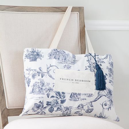 White and blue patterned tote bag with "French Bedroom" text, hanging on a beige chair. Features a blue tassel accent.
